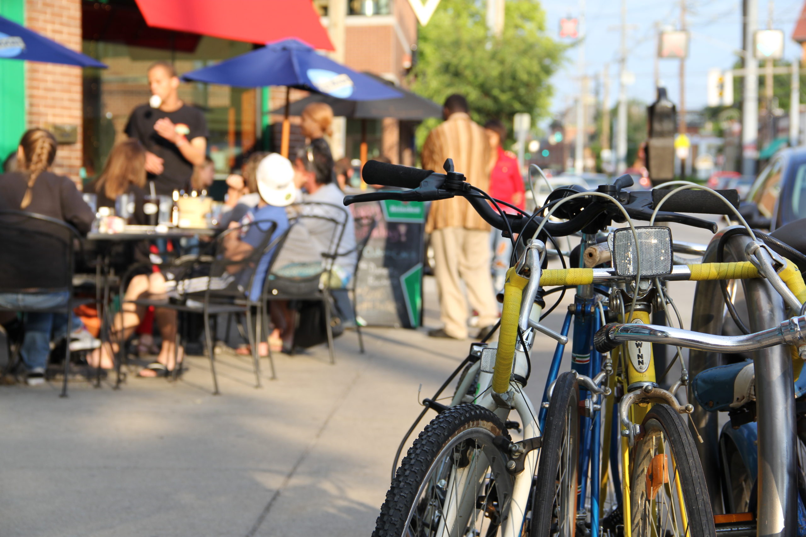 A yellow bike is tied to a bike stand with people eating brunch on a patio on the background.